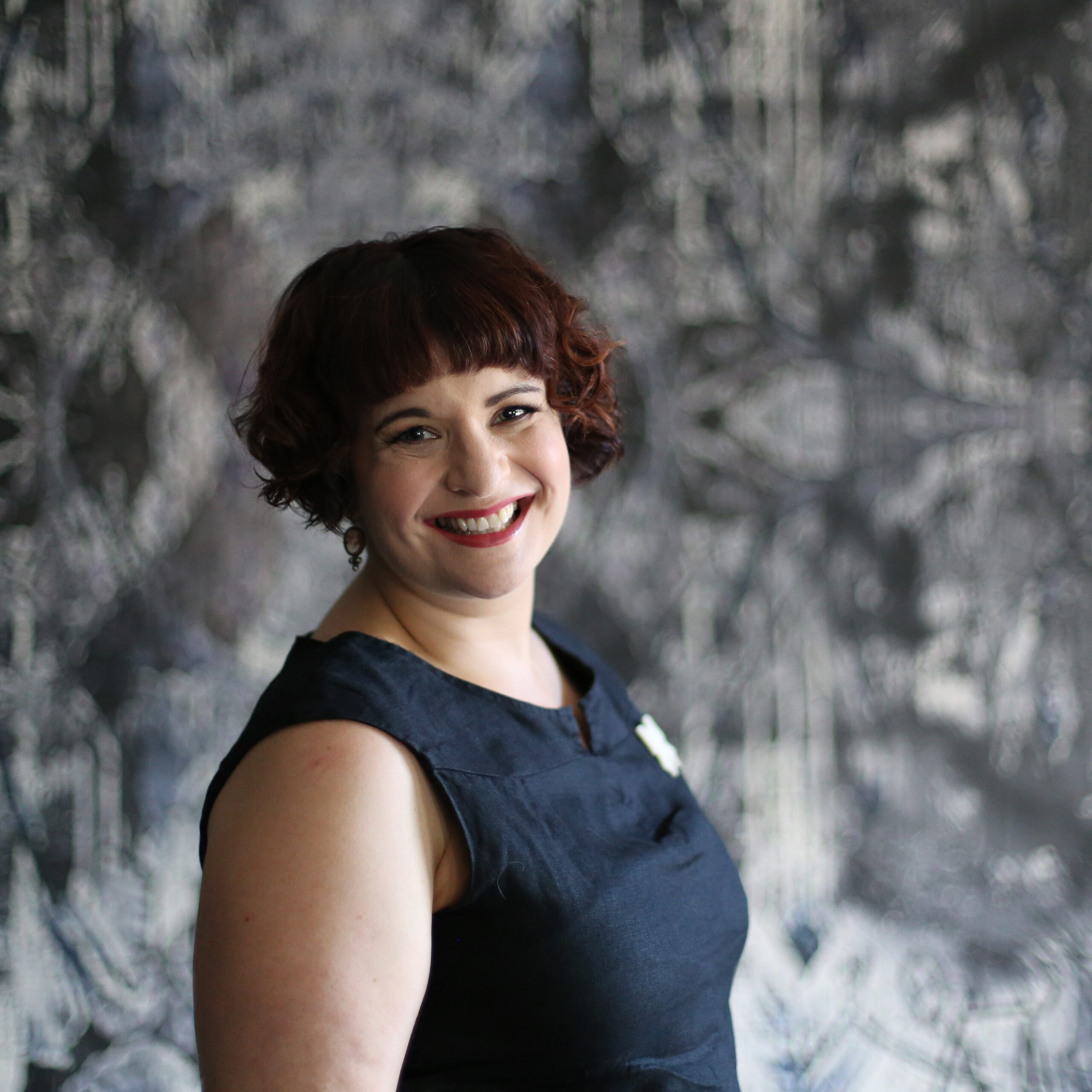 Kate Schirmer, a woman with short curly hair and wearing red lipstick, smiling broadly.