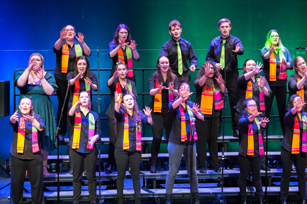 Three rows of performers wearing colourful scarves while standing on choir risers. They are reaching their arms out in front of them.