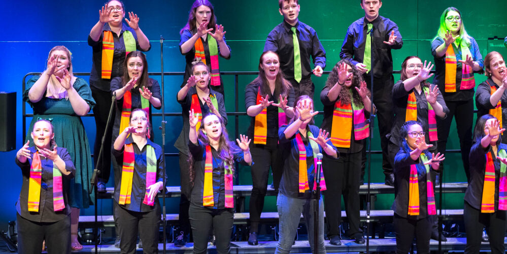 Three rows of performers wearing colourful scarves while standing on choir risers. They are reaching their arms out in front of them.