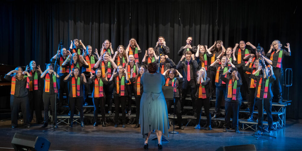 Three rows of people wearing colourful scarves while standing on choir risers. They are all clutching the sides of their heads with their hands.