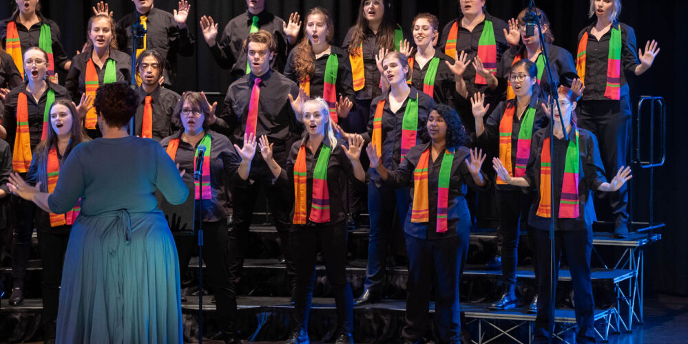 Three rows of people wearing colourful scarves standing on choir risers. They all have their hands raised beside their heads with their palms facing outwards.